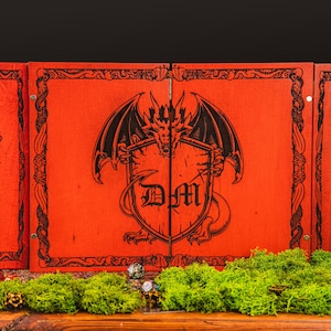 May include: A three-panel, red tabletop gaming screen with a black dragon and shield design. The shield has the letters "DM" on it. The screen is decorated with a black border and hinges. Green moss and dice are in the foreground.