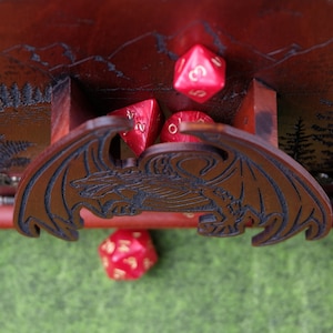 May include: A dark brown wooden dice tower featuring a laser-engraved dragon on the front panel and a mountain forest scene on the back. The tower includes a small tray area containing several translucent red multi-sided gaming dice.