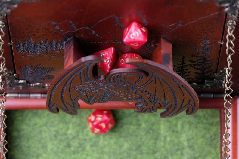 May include: A dark brown wooden dice tower featuring a laser-engraved dragon on the front panel and a mountain forest scene on the back. The tower includes a small tray area containing several translucent red multi-sided gaming dice.