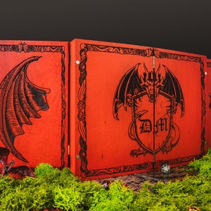 May include: A red wooden tabletop game screen with black dragon wing and shield designs. The center panel features a dragon emblem with the letters "DM". The screen is set on a bed of green moss.