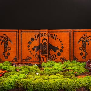 Peut inclure: Un &eacute;cran de ma&icirc;tre de donjon orange avec des illustrations et du texte noirs. L'&eacute;cran est entour&eacute; de mousse verte, avec un bougeoir dragon et d'autres accessoires sur le th&egrave;me de la fantaisie.