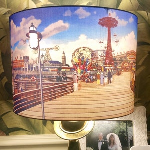 May include: A vintage-style table lamp with a black metal base and a gold-colored shade. The shade features a colorful illustration of a boardwalk amusement park with a Ferris wheel, a roller coaster, and a carousel. The lamp is sitting on a wooden table.