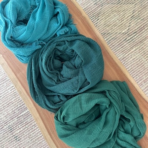 May include: Three teal and green scarves are arranged on a wooden tray. The scarves are made of a soft, textured fabric and have a fringed edge. The scarves are in shades of teal, dark teal, and green.