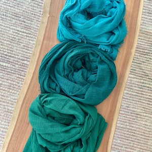 May include: Three teal, green, and emerald green scarves are arranged on a wooden surface. The scarves are made of a soft, lightweight fabric and have a fringed edge. The scarves are neatly folded and appear ready to wear.