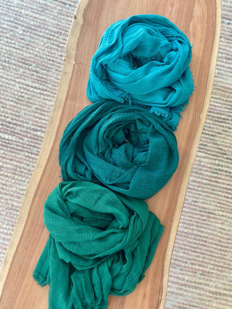 May include: Three teal, green, and emerald green scarves are arranged on a wooden surface. The scarves are made of a soft, lightweight fabric and have a fringed edge. The scarves are neatly folded and appear ready to wear.