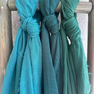 May include: Three knotted scarves in shades of teal and green hang from a wooden rod. The scarves have a textured appearance and frayed edges. The colors range from a bright teal to a deep forest green.