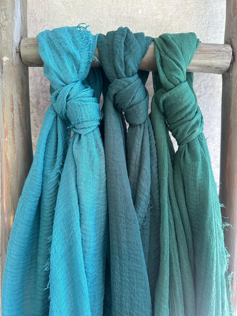 May include: Three knotted scarves in shades of teal and green hang from a wooden rod. The scarves have a textured appearance and frayed edges. The colors range from a bright teal to a deep forest green.