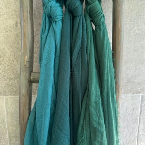May include: Three teal and green scarves are knotted and draped over a rustic wooden ladder. The scarves vary in shades from light teal to deep green, with a textured, fringed edge. The ladder is weathered, adding a vintage aesthetic.
