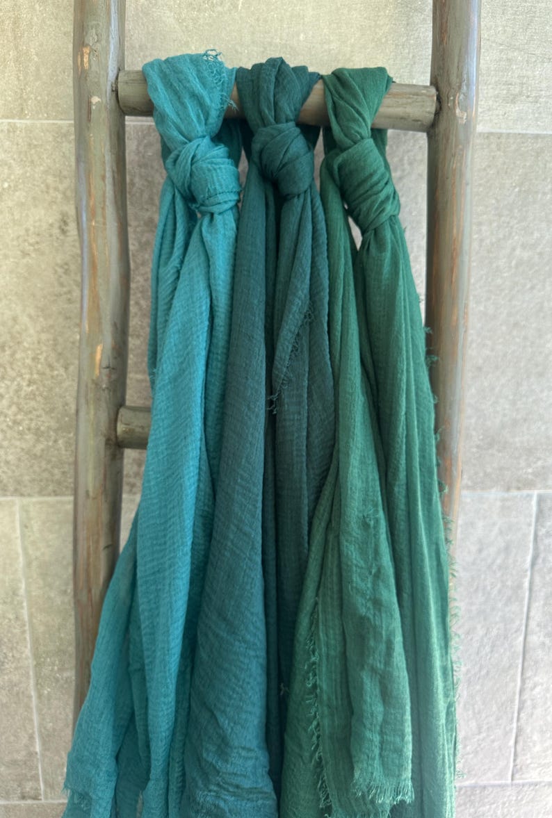 May include: Three teal and green scarves are knotted and draped over a rustic wooden ladder. The scarves vary in shades from light teal to deep green, with a textured, fringed edge. The ladder is weathered, adding a vintage aesthetic.