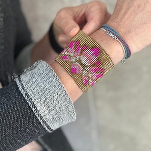 May include: A gold beaded bracelet with a pink and white floral design. The bracelet is worn on a person's wrist with a second bracelet that says "MAMA" in silver letters.
