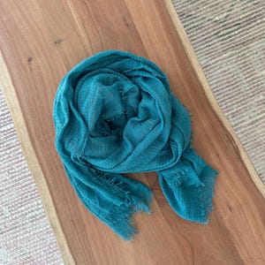May include: A teal-colored scarf with a fringed edge is draped on a wooden surface. The scarf is made of a lightweight fabric and has a slightly wrinkled texture. The wooden surface has a natural wood grain.