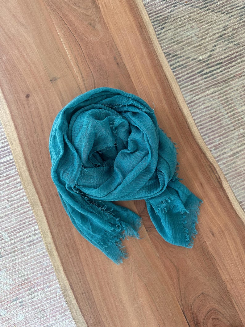 May include: A teal-colored scarf with a fringed edge is draped on a wooden surface. The scarf is made of a lightweight fabric and has a slightly wrinkled texture. The wooden surface has a natural wood grain.