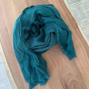 May include: A teal-colored scarf with a fringed edge, draped on a wooden surface. The scarf is made of a lightweight fabric, with a slightly textured appearance. The scarf is a solid color and appears to be new.