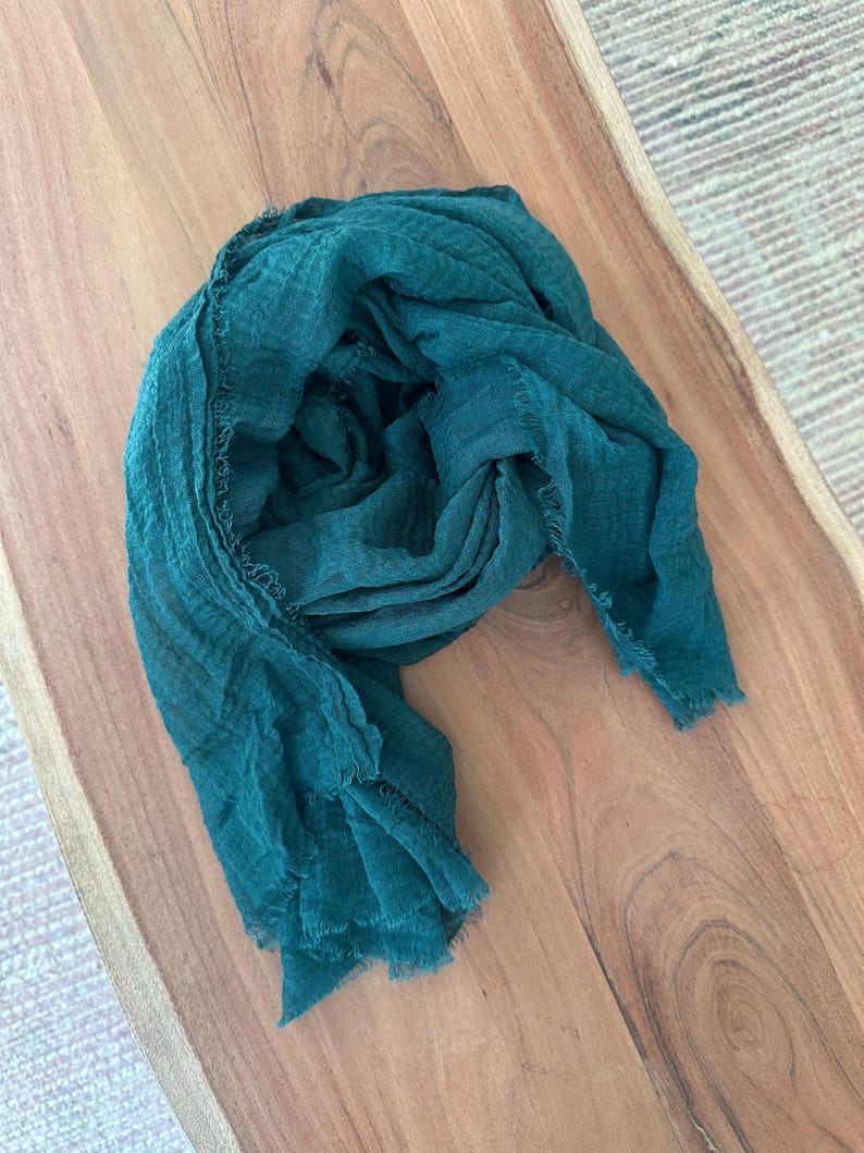 May include: A teal-colored scarf with a fringed edge, draped on a wooden surface. The scarf is made of a lightweight fabric, with a slightly textured appearance. The scarf is a solid color and appears to be new.