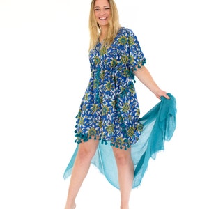 May include: A blue floral print dress with a V-neck and tassel trim. The dress has a drawstring waist and is paired with a flowing teal scarf. The dress is knee-length and has short sleeves. The model is smiling.