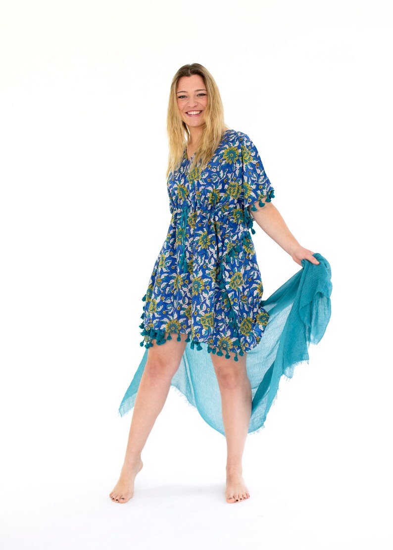 May include: A blue floral print dress with a V-neck and tassel trim. The dress has a drawstring waist and is paired with a flowing teal scarf. The dress is knee-length and has short sleeves. The model is smiling.