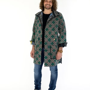 May include: A man wearing a long, black and green patterned coat with a black collar and a gold button. He is also wearing blue jeans and brown shoes.