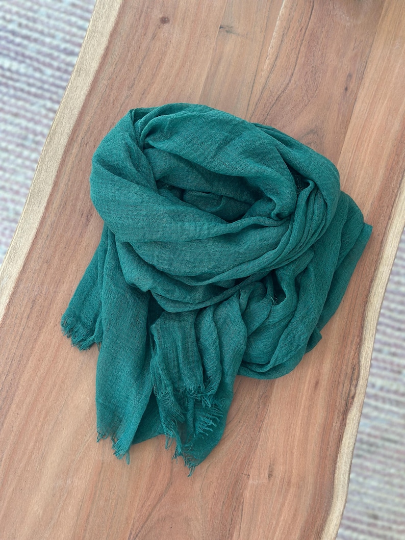 May include: A teal-colored scarf made of a lightweight fabric, draped on a wooden surface. The scarf has a fringed edge and a slightly textured appearance. The scarf is a solid color and appears to be soft and flowing.