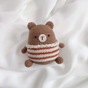 May include: A brown crocheted bear wearing a white and orange striped shirt. The bear has black eyes and a brown nose.