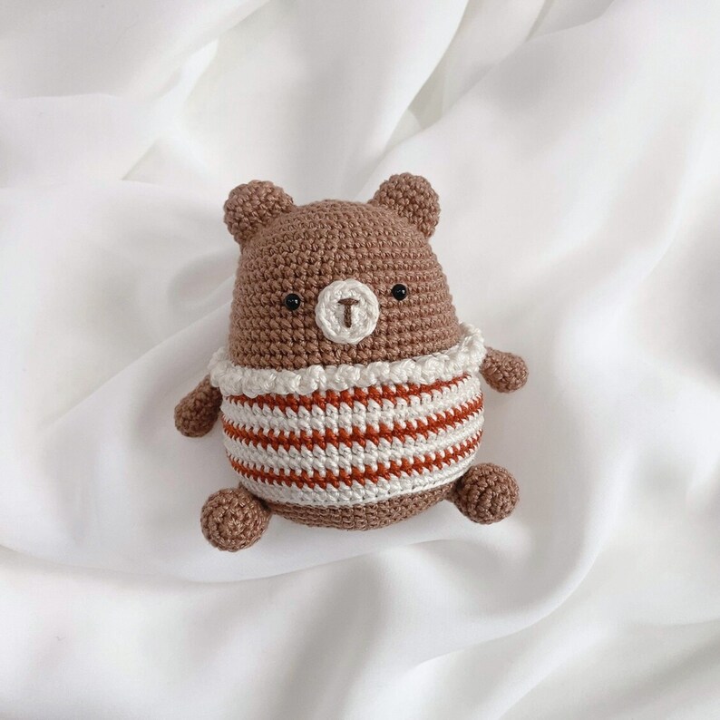 May include: A brown crocheted bear wearing a white and orange striped shirt. The bear has black eyes and a brown nose.