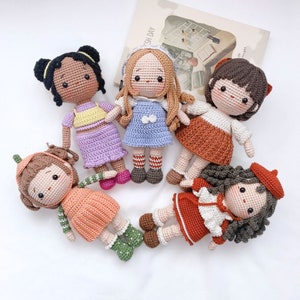 May include: Five crocheted dolls with different colored dresses and hair. The dolls are all standing on a white surface.