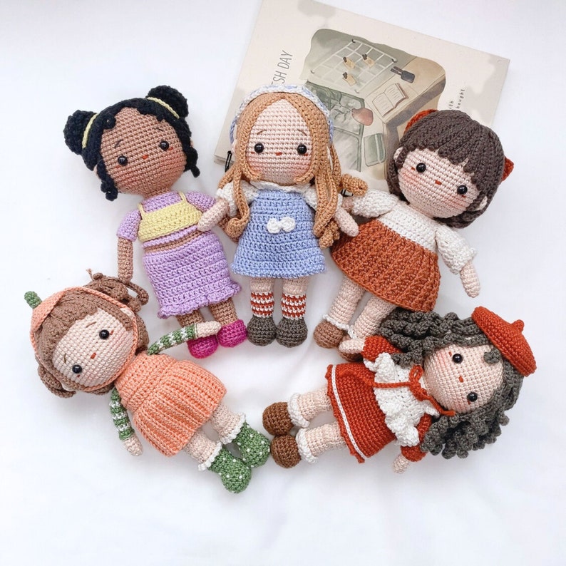 May include: Five crocheted dolls with different colored dresses and hair. The dolls are all standing on a white surface.