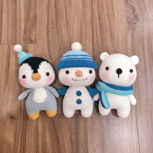 May include: Three crocheted stuffed animals: a penguin, a snowman, and a polar bear. The penguin is blue and gray, the snowman is white with a blue hat, and the polar bear is white with a blue scarf.