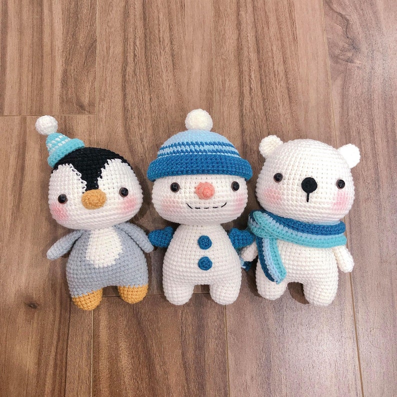 May include: Three crocheted stuffed animals: a penguin, a snowman, and a polar bear. The penguin is blue and gray, the snowman is white with a blue hat, and the polar bear is white with a blue scarf.