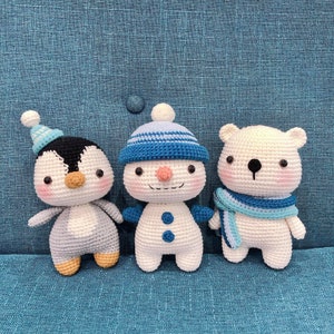 May include: Three crocheted stuffed animals: a penguin, a snowman, and a polar bear. The penguin is wearing a blue hat, the snowman is wearing a blue and white striped hat, and the polar bear is wearing a blue scarf. All three animals are white with black eyes and black noses.