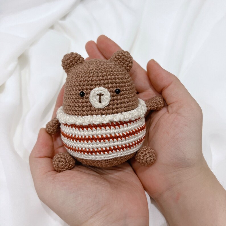 May include: A brown crocheted bear wearing a red and white striped sweater. The bear has black eyes and a brown nose.