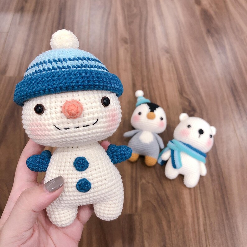 May include: A crocheted snowman with a blue and white hat, a crocheted penguin with a blue hat, and a crocheted polar bear with a blue scarf.