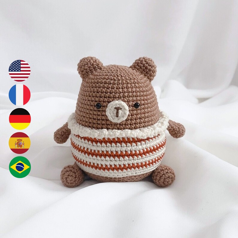 May include: A brown crocheted bear wearing a white and orange striped shirt. The bear has black eyes and a brown nose.