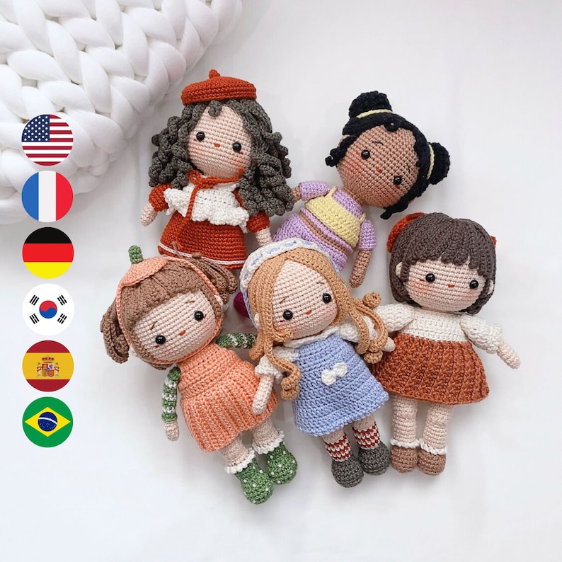 May include: Five crocheted dolls with different colored dresses and hair. The dolls are all smiling and have brown eyes. The dolls are sitting on a white surface.