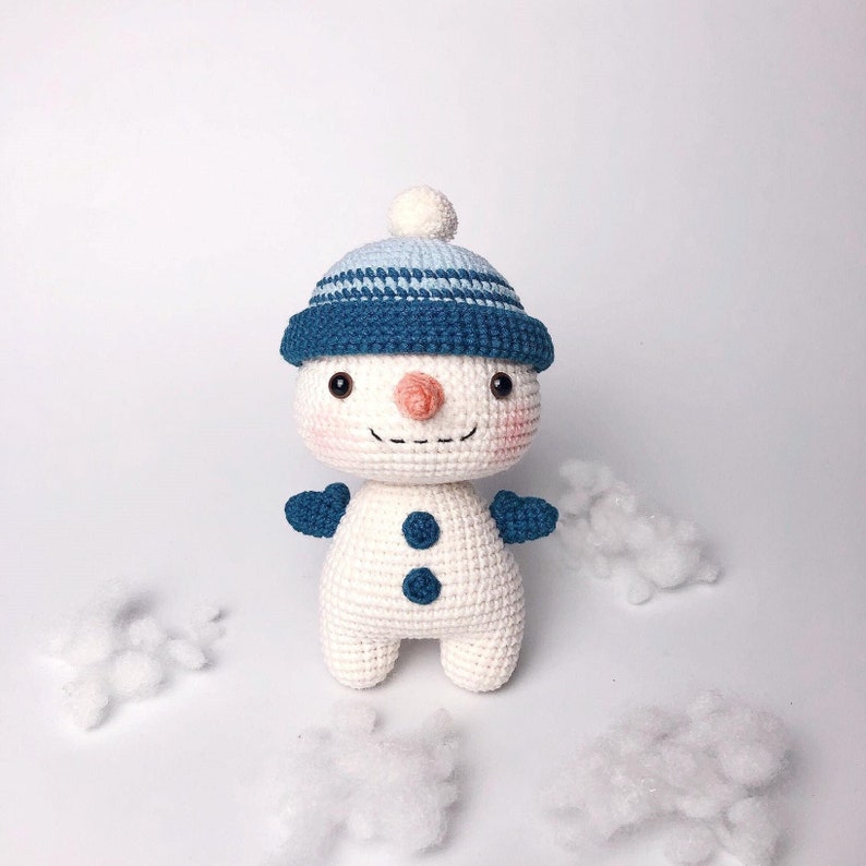 May include: A white crocheted snowman with a blue and white striped hat and blue mittens. The snowman has a carrot nose, black eyes, and a smiling mouth.