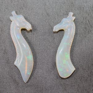 May include: Two giraffe-shaped opal pendants. The pendants are carved with a curved neck and detailed head. The opals have a milky white base color with flashes of iridescent green and blue. The pendants are displayed on a gray textured surface.