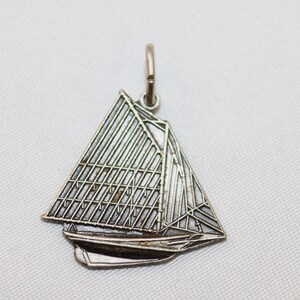 May include: A silver-toned sailboat pendant with a detailed design. The pendant features a triangular sail with a grid pattern and a curved hull. The bail is a small loop at the top. The pendant is likely made of metal and is suitable for a necklace or bracelet.