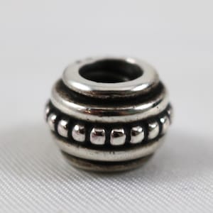May include: A silver bead with a cylindrical shape. The bead features a series of horizontal ridges and a row of small, round accents. The top has a large hole, and the bead is set against a white background.