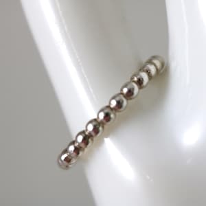 May include: A silver beaded ring with a row of small, round beads. The ring is shown against a white background, highlighting its metallic shine and simple design. The beads are evenly spaced, creating a delicate and minimalist aesthetic.