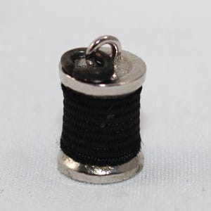 May include: A small, cylindrical metal charm shaped like a spool of thread, featuring a shiny silver-toned finish on the top and bottom ends and wrapped with black thread around the center, topped with a small silver-toned metal jump ring.