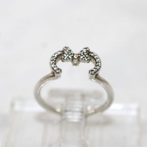 May include: A silver ring featuring a bow design. The bow is adorned with small, sparkling stones. The ring has a delicate, open design, with the bow as the central focus. The ring is displayed on a clear surface.