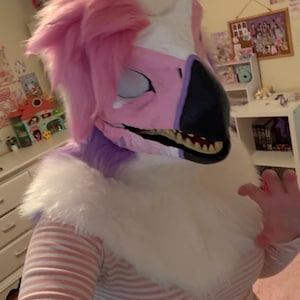 May include: A pink, white, and black fursuit head with a feathery mane. The fursuit has a beak and visible teeth. The person wearing the fursuit is also wearing a pink and white striped shirt.
