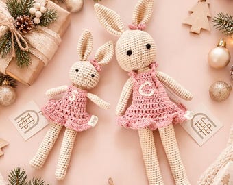 May include: Two cream-colored crocheted rabbit dolls with long ears, wearing pink dresses with the letters S and C embroidered on the chests. Each doll has a small tag attached that reads TEP TEP. The dolls are surrounded by holiday decorations.