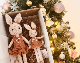 May include: Two crocheted bunny toys in a gift box. The larger bunny is wearing a brown dress with a white "C" on the front. The smaller bunny is wearing a brown dress with a white "M" on the front. The bunnies are sitting on brown packing material.