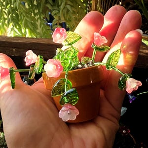 以下が含まれることがあります： 半透明のピンクのガラスの花と鮮やかな緑のガラスの葉が細いワイヤーの茎についたミニチュアの鉢植え。植物は小さな茶色のテラコッタ色の鉢に入っており、繊細で詳細な植物のディスプレイを形成しています。