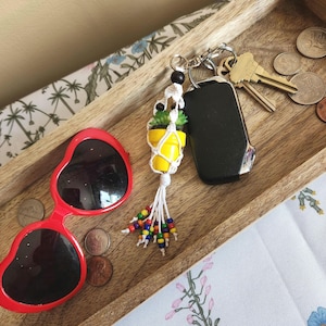 May include: A white macrame keychain with a small yellow and green potted plant charm. The keychain has colourful beads and is attached to a silver keyring with keys.
