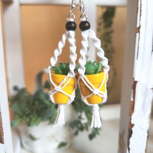 May include: A pair of yellow potted plant earrings with white macrame hangers and brown wooden beads. The earrings are hanging from a white surface.