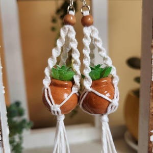 May include: A pair of white macrame earrings with small terracotta pots and green plants hanging from them. The pots have a brown wooden bead at the top.