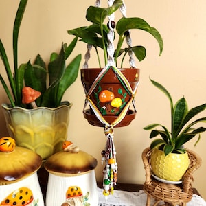 Puede incluir: Un macramé para colgar plantas con una maceta marrón y una planta verde. La maceta está decorada con setas pintadas. El macramé tiene un borla marrón y blanco. Hay dos jarras de cerámica con forma de seta en el fondo.