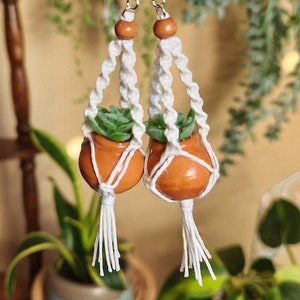 May include: A pair of handmade earrings featuring miniature terracotta pots with green plants hanging from white macrame cords. The earrings have silver hooks.