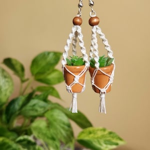 May include: A pair of white macrame hanging planter earrings with small terracotta pots and green plants. The earrings have wooden beads and silver hooks.